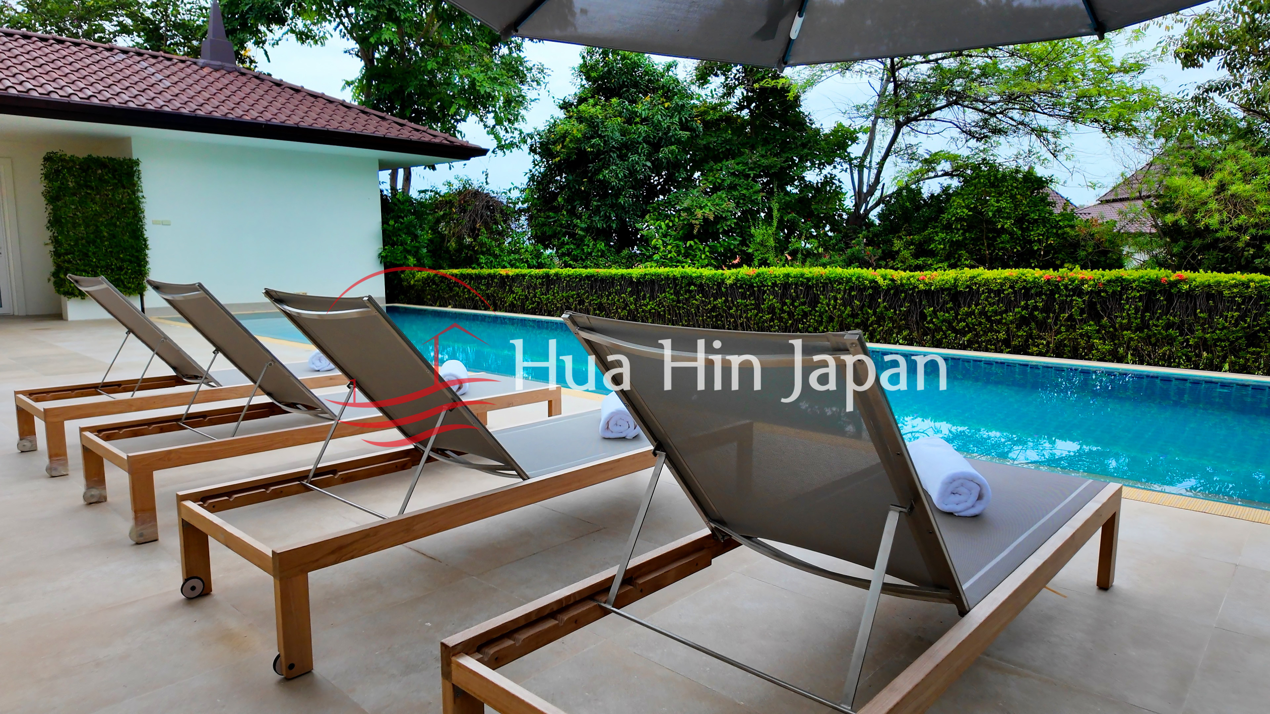 Luxury outdoor swimming pool with wooden sun loungers, rolled towels, tropical landscaping, and privacy hedges