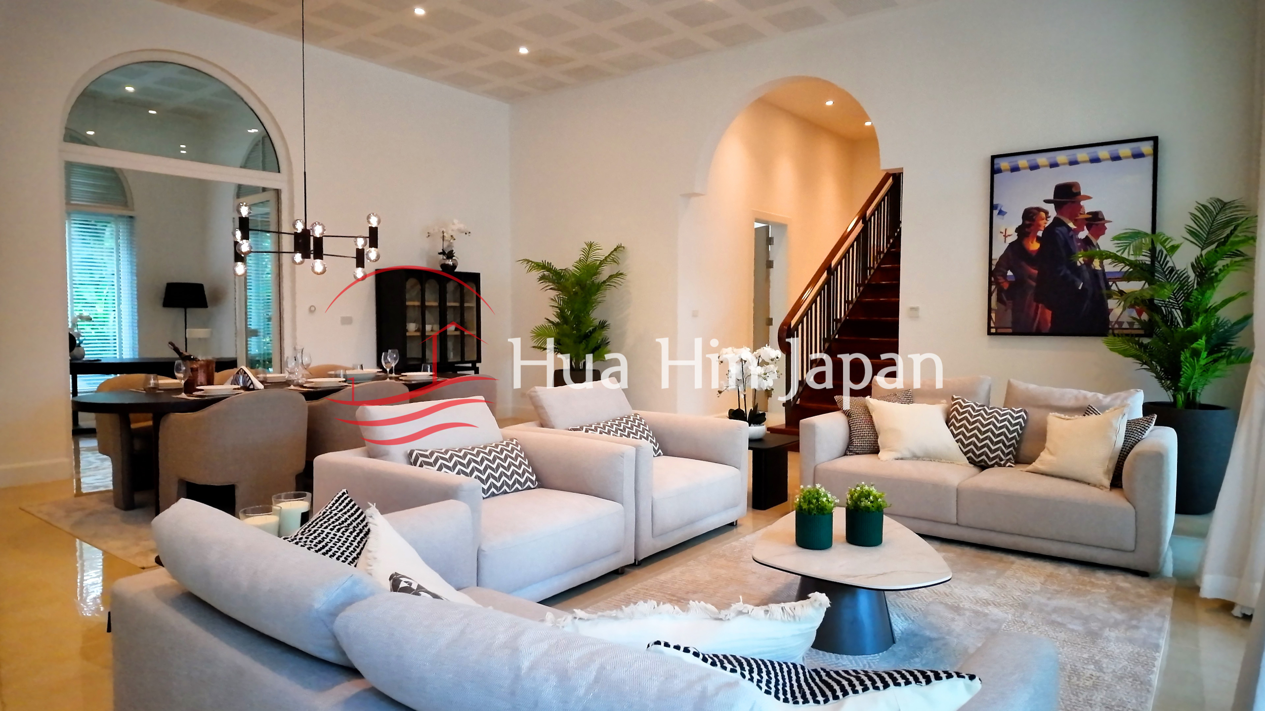 Elegant open-plan living room with cream sofas, patterned cushions, modern chandelier, indoor plants, artwork, and wooden staircase