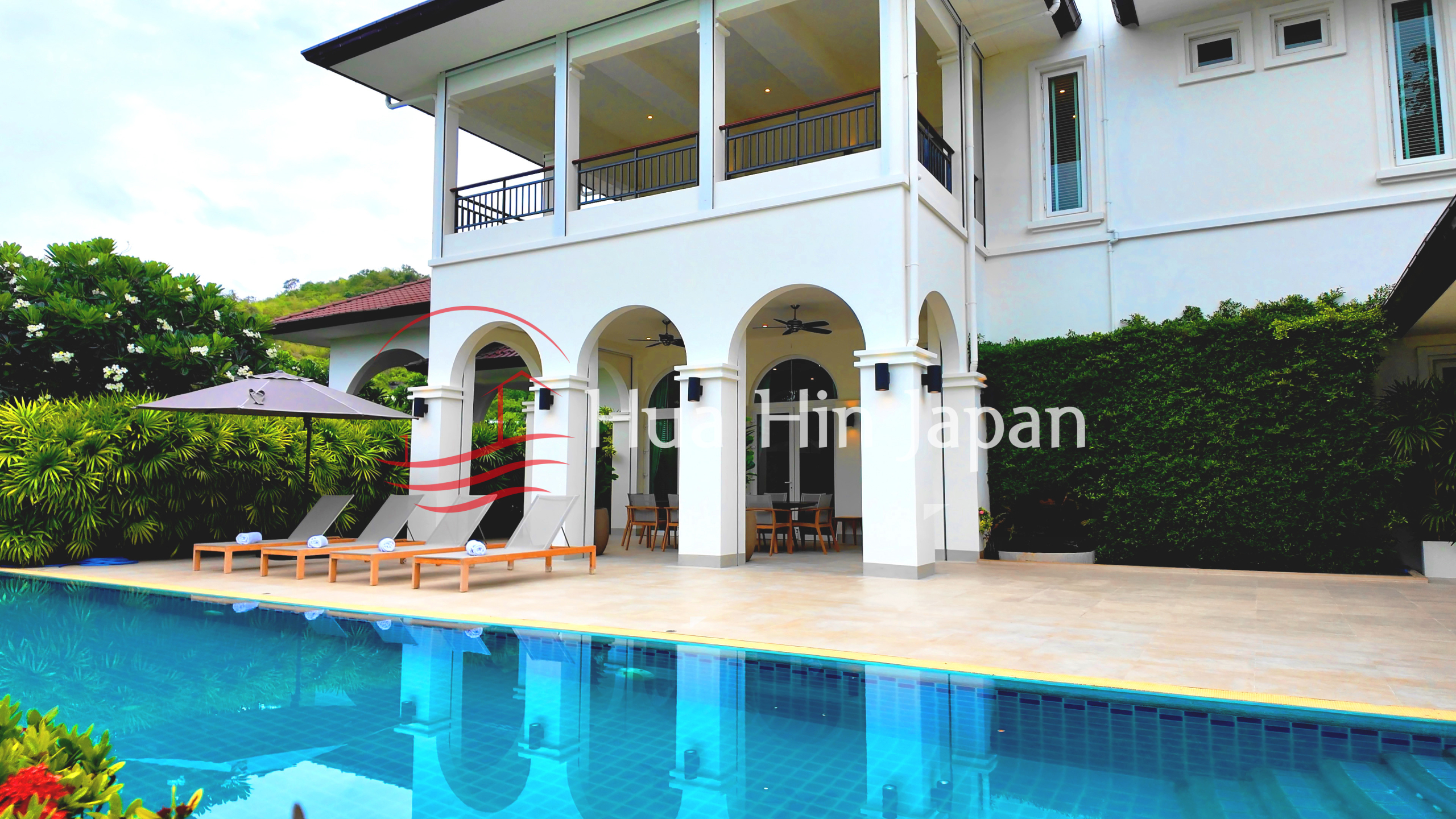 Luxury villa with a large swimming pool, outdoor lounge chairs, arched terrace, and lush tropical landscaping