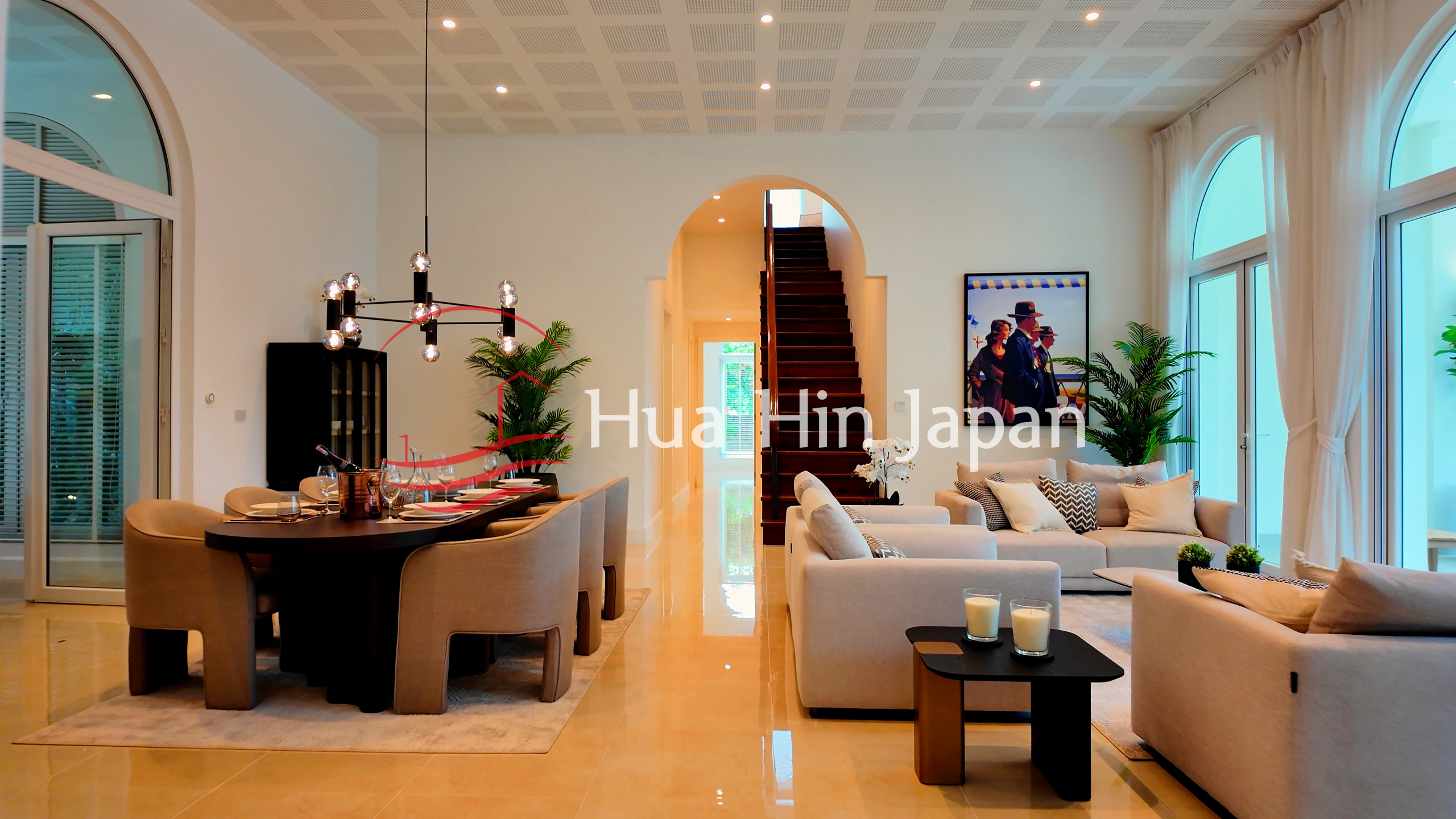 Elegant open-plan living and dining room with cream sofas, a round dining table, arched windows, chandelier, and staircase