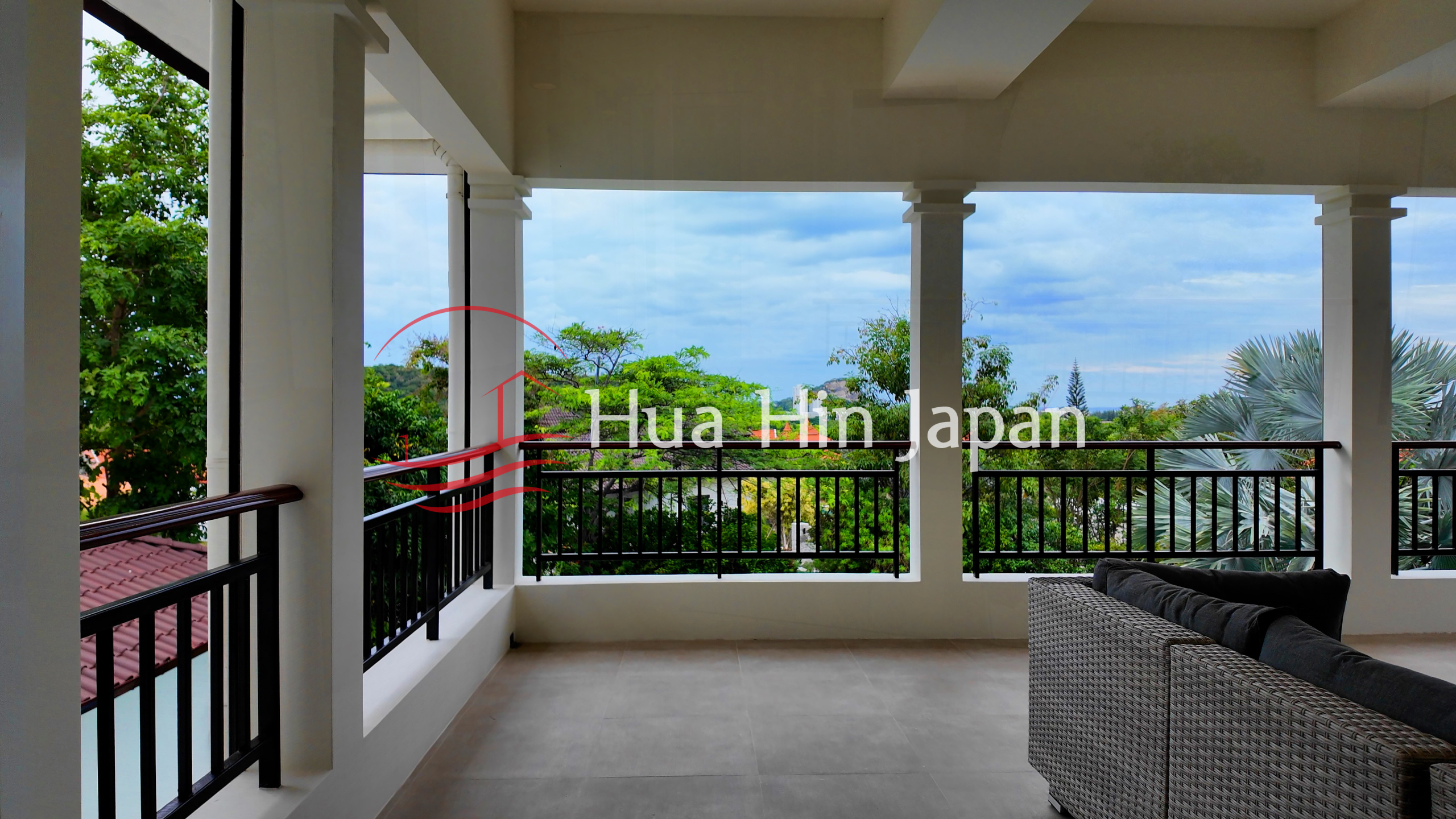 Spacious covered balcony with outdoor seating, white columns, black railings, and lush garden views