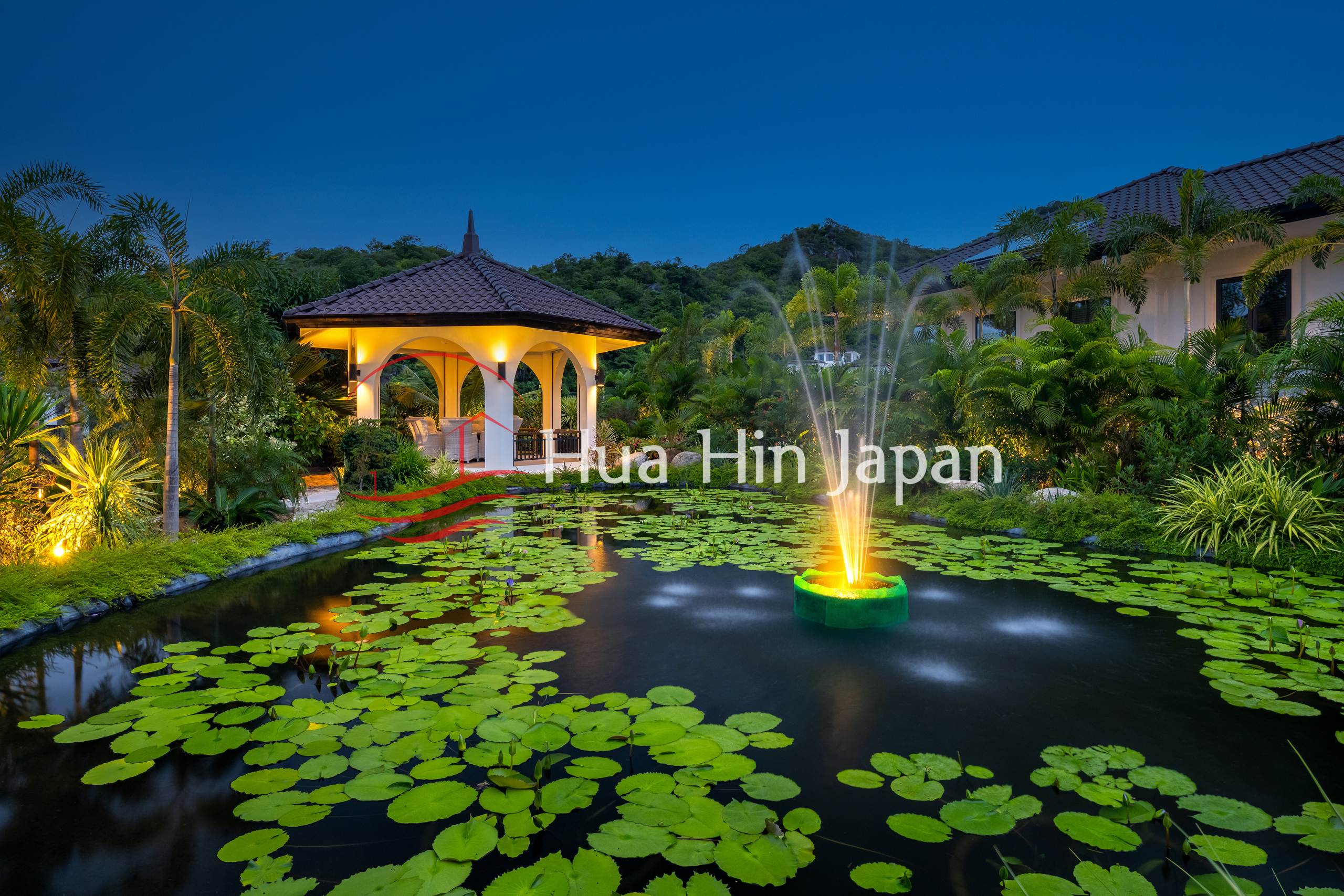 Tropical resort garden with a lotus pond, fountain, and illuminated pavilion at dusk.