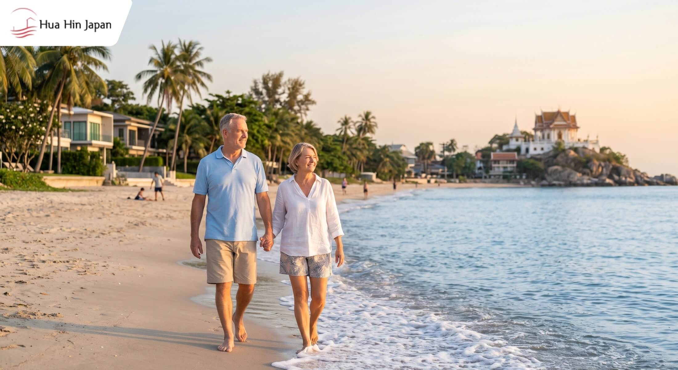 A senior expat couple walking along the sandy beach in Hua Hin, Thailand, during sunset with luxury villas and a Thai temple in the background.