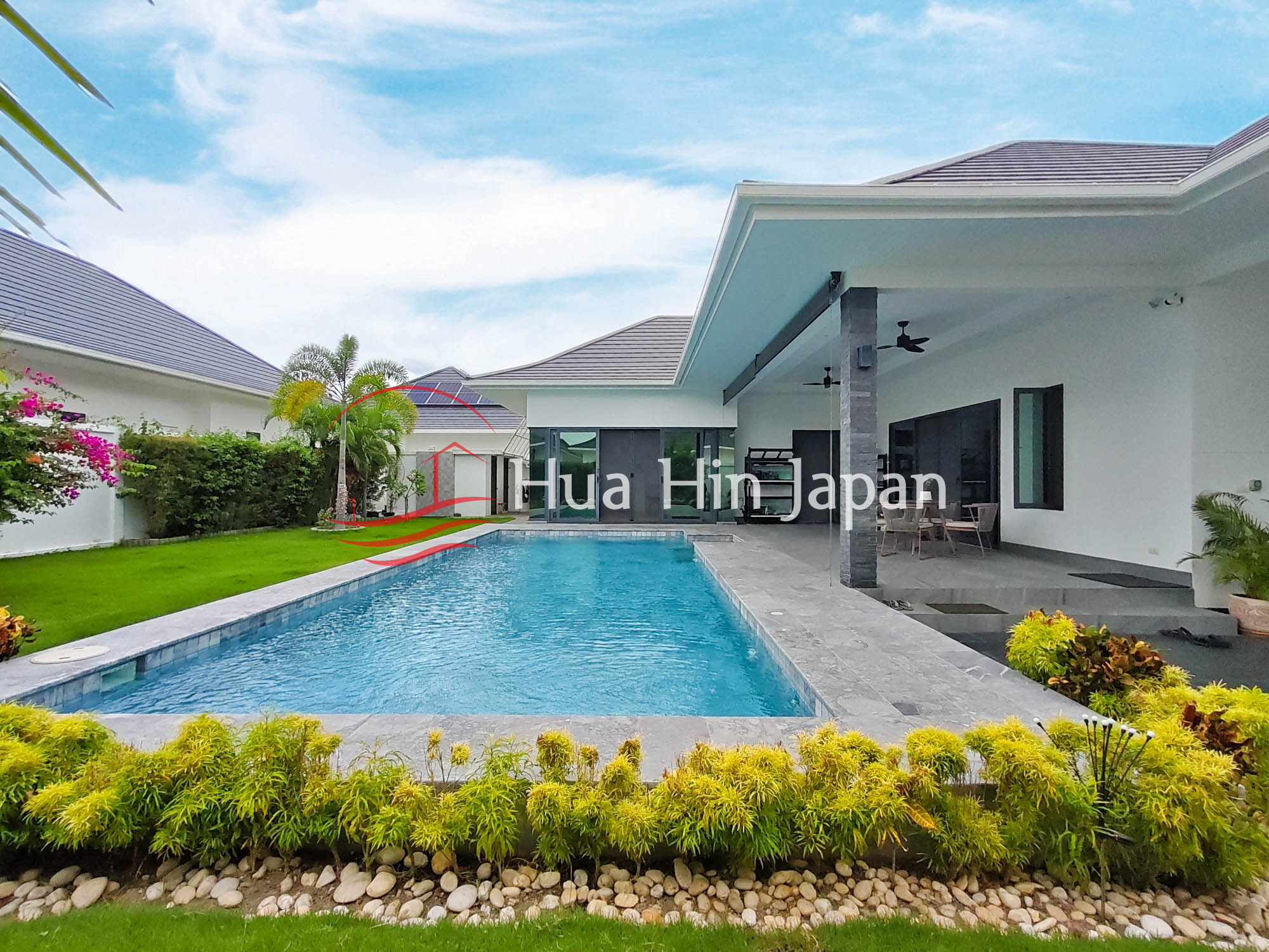 Modern white pool villa with a large rectangular swimming pool, covered patio, green lawn, and landscaped garden