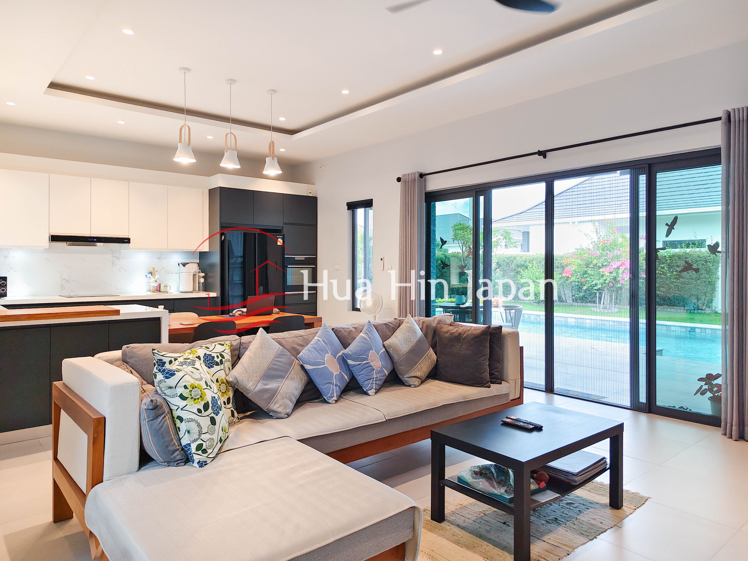 Bright modern living room with a cream sectional sofa, patterned cushions, black coffee table, open kitchen, and large glass doors overlooking a swimming pool