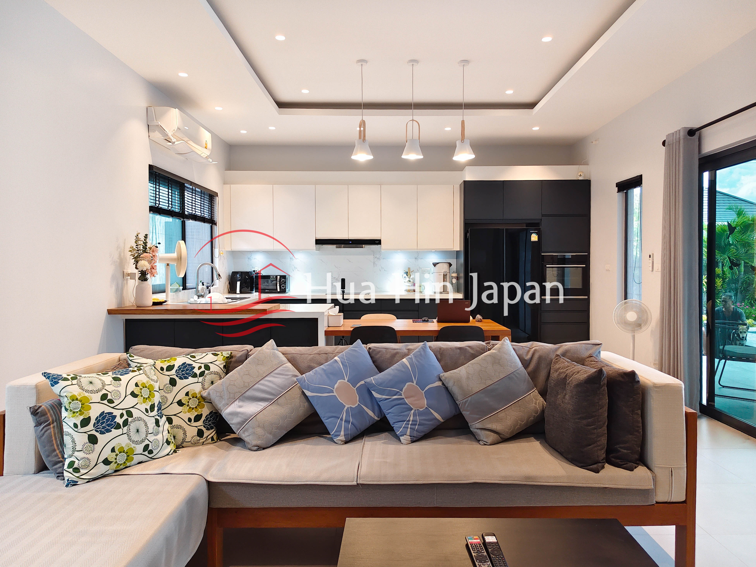 Bright open-plan living room and kitchen with a large sofa, patterned cushions, dining table, modern black and white kitchen cabinets, and large windows