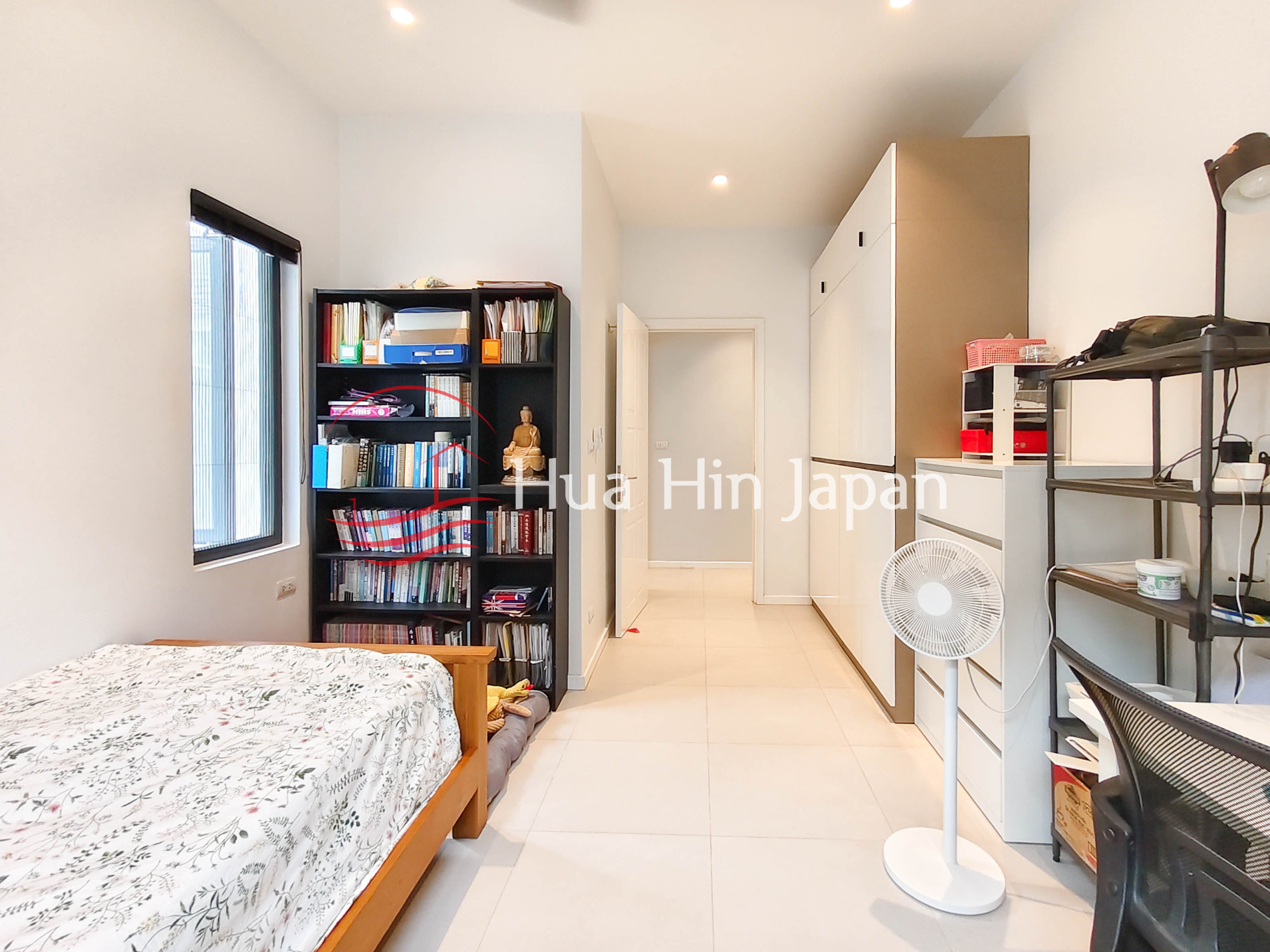 Bright bedroom with wooden bed, large bookshelf, built-in wardrobes and standing fan