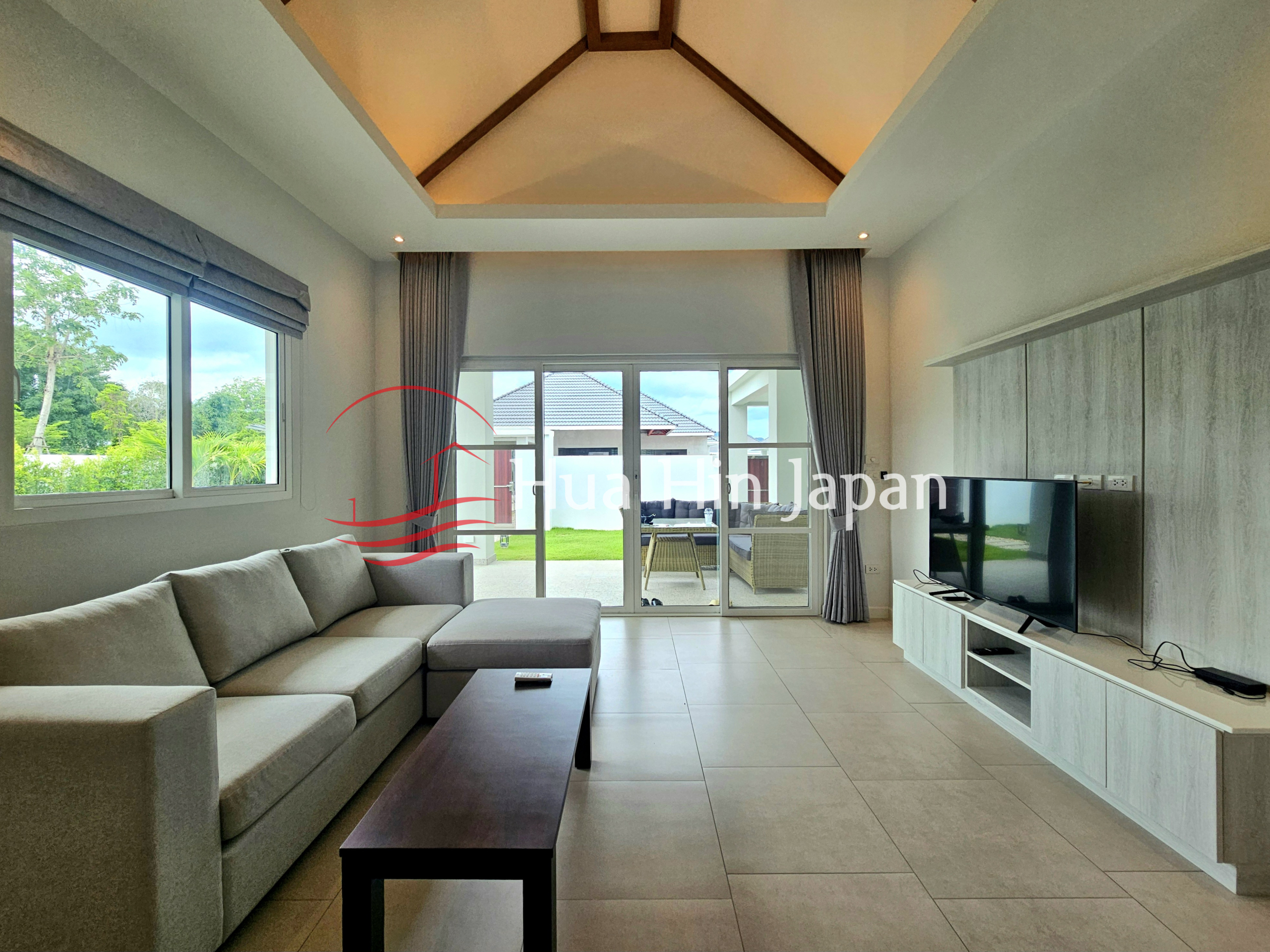 Spacious modern living room with an L-shaped sofa, wooden coffee table, large TV, and glass doors opening to a green outdoor area