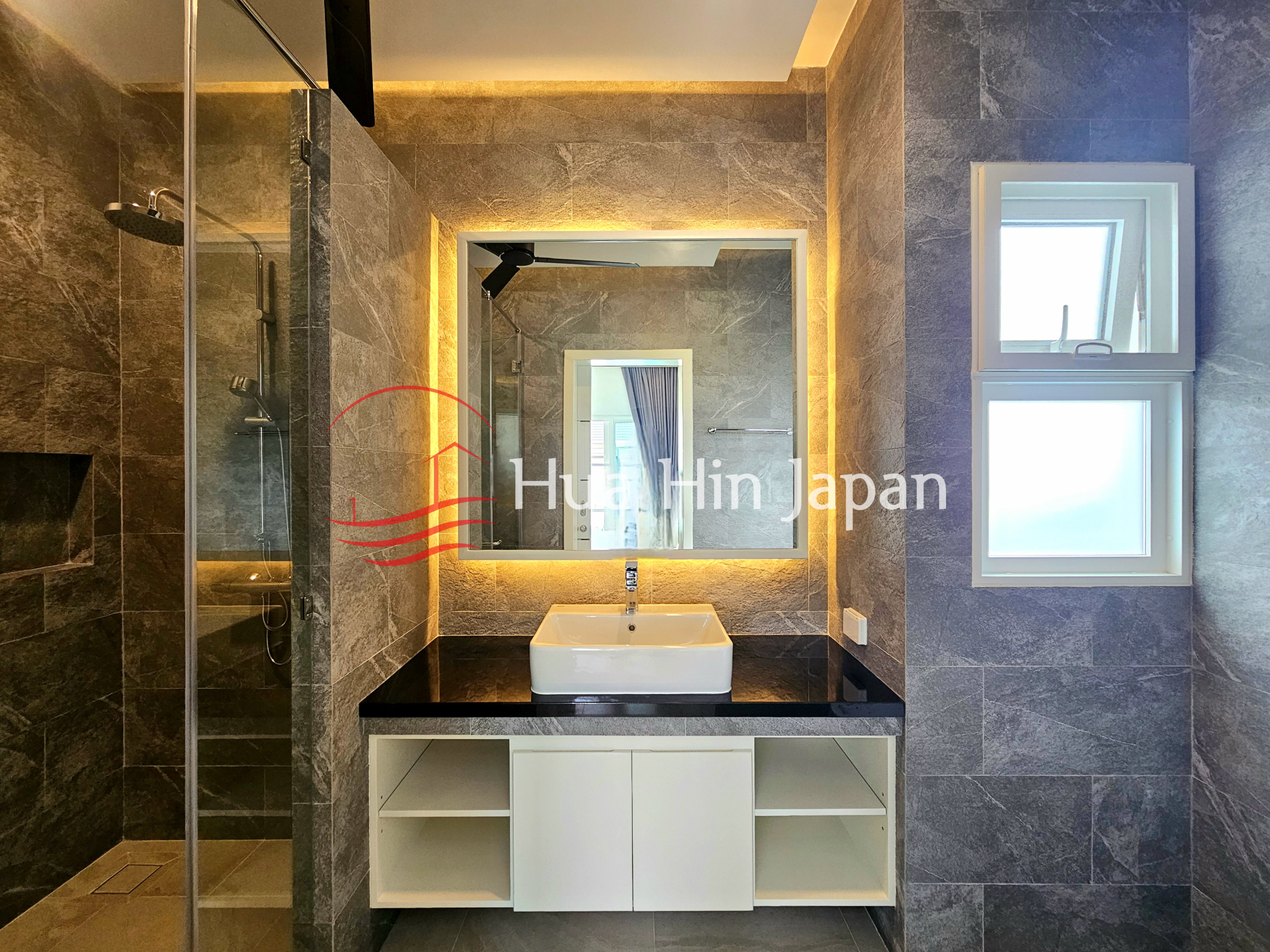 Modern bathroom with stone-look tiles, glass shower enclosure, white vanity, illuminated mirror, and frosted windows