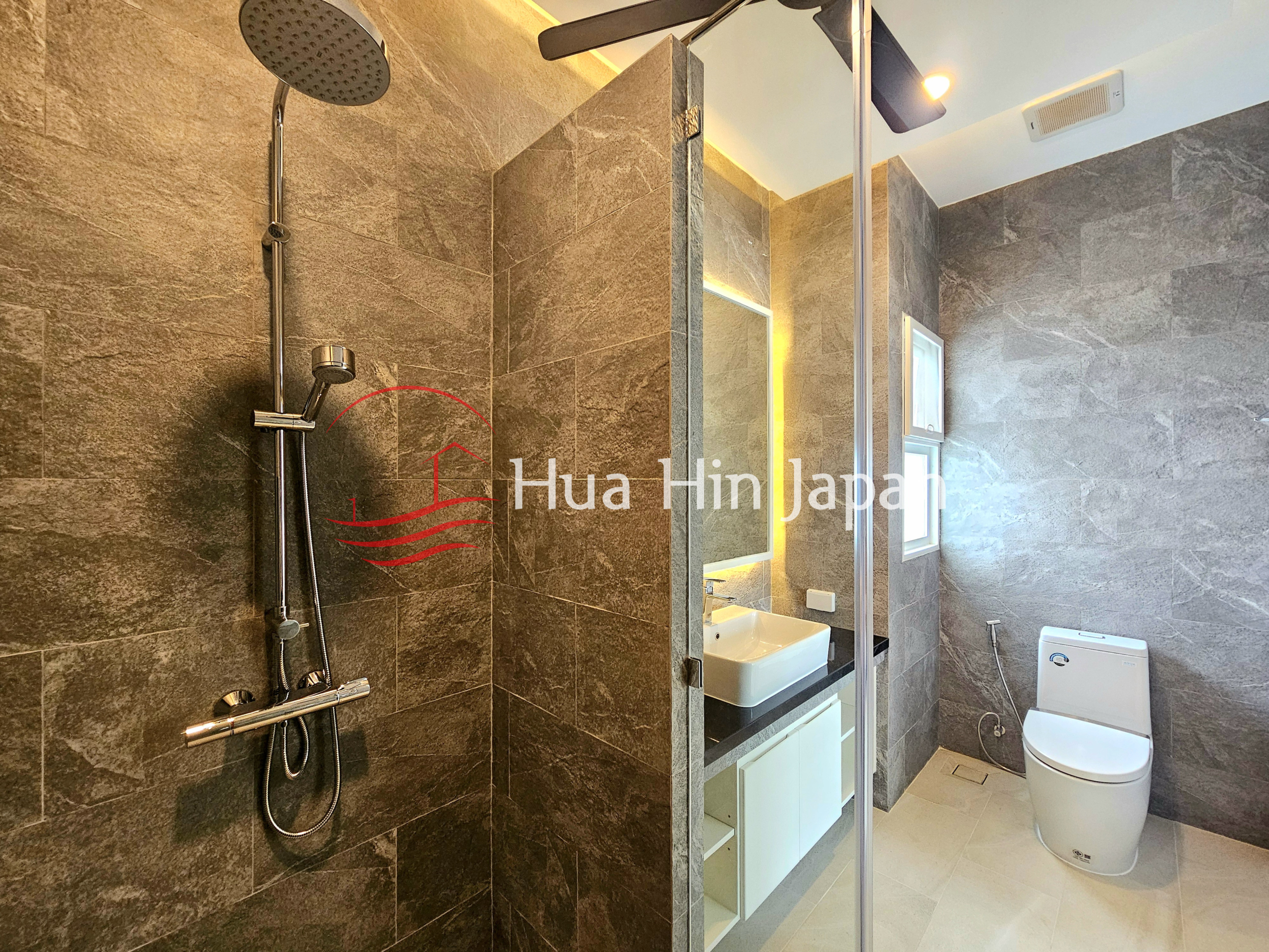 Modern bathroom with stone-look tiles, glass shower enclosure, rainfall shower, white toilet, vanity sink, and illuminated mirror