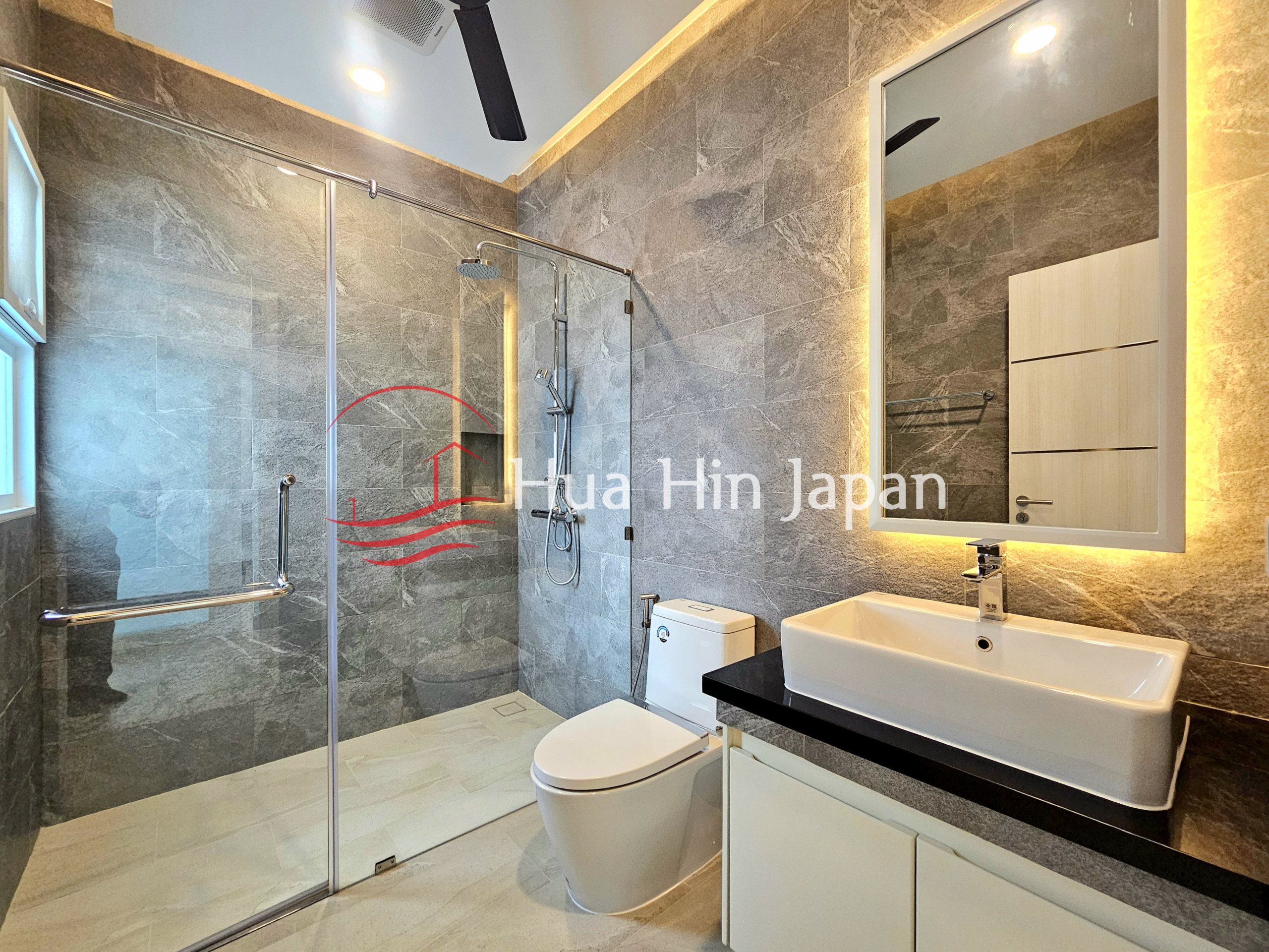Modern bathroom with gray stone-look tiles, large glass shower, white toilet, vanity sink, and illuminated mirror