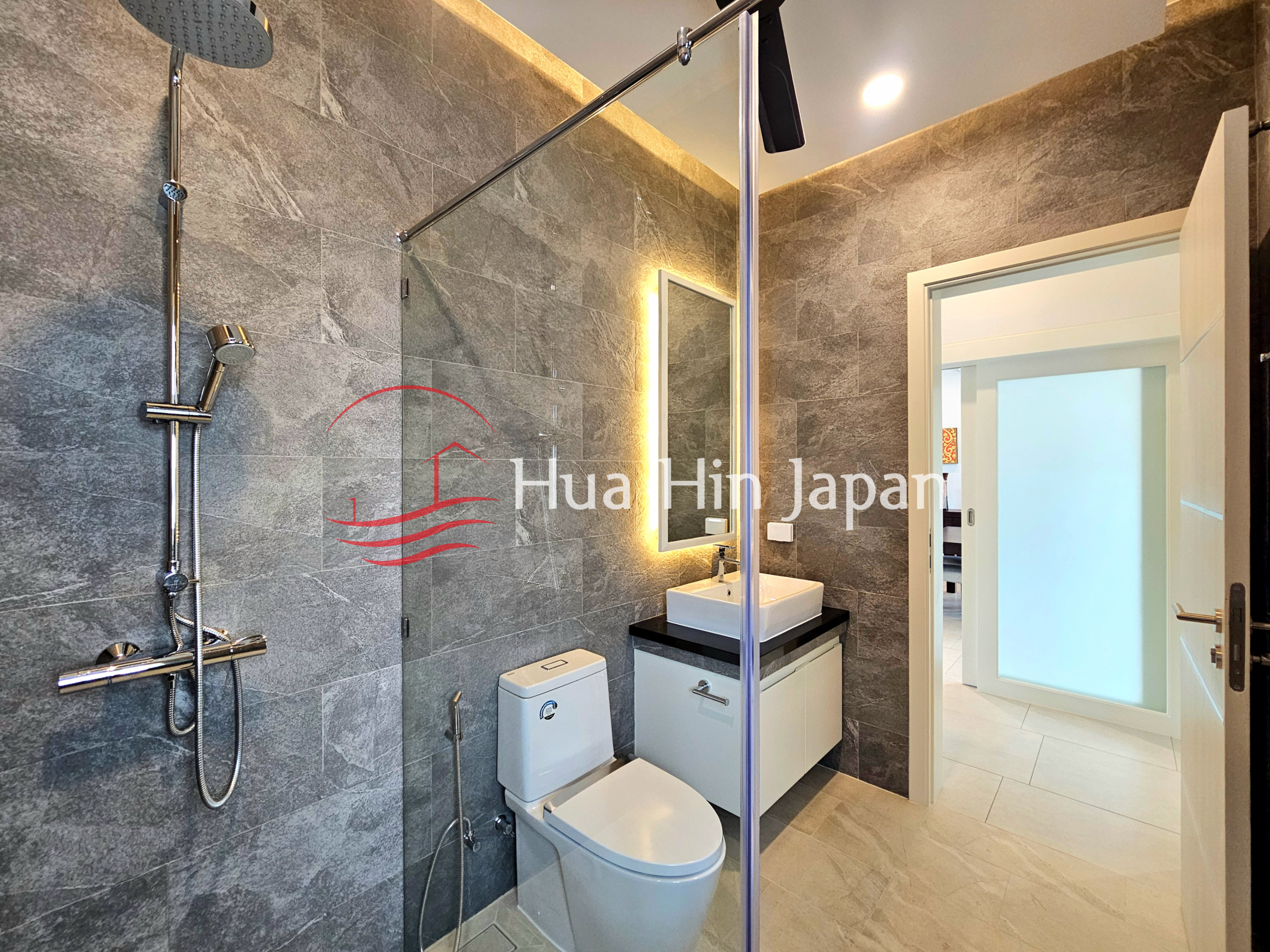 Modern bathroom with gray stone-look tiles, glass shower enclosure, wall-mounted shower, toilet, vanity, and illuminated mirror