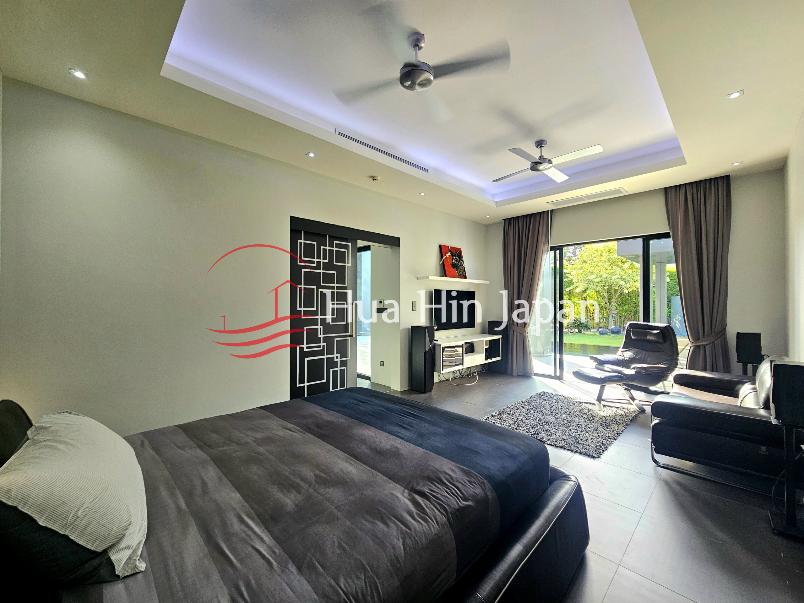 Spacious modern bedroom and living area with a large bed, leather seating, ceiling fans, and garden view