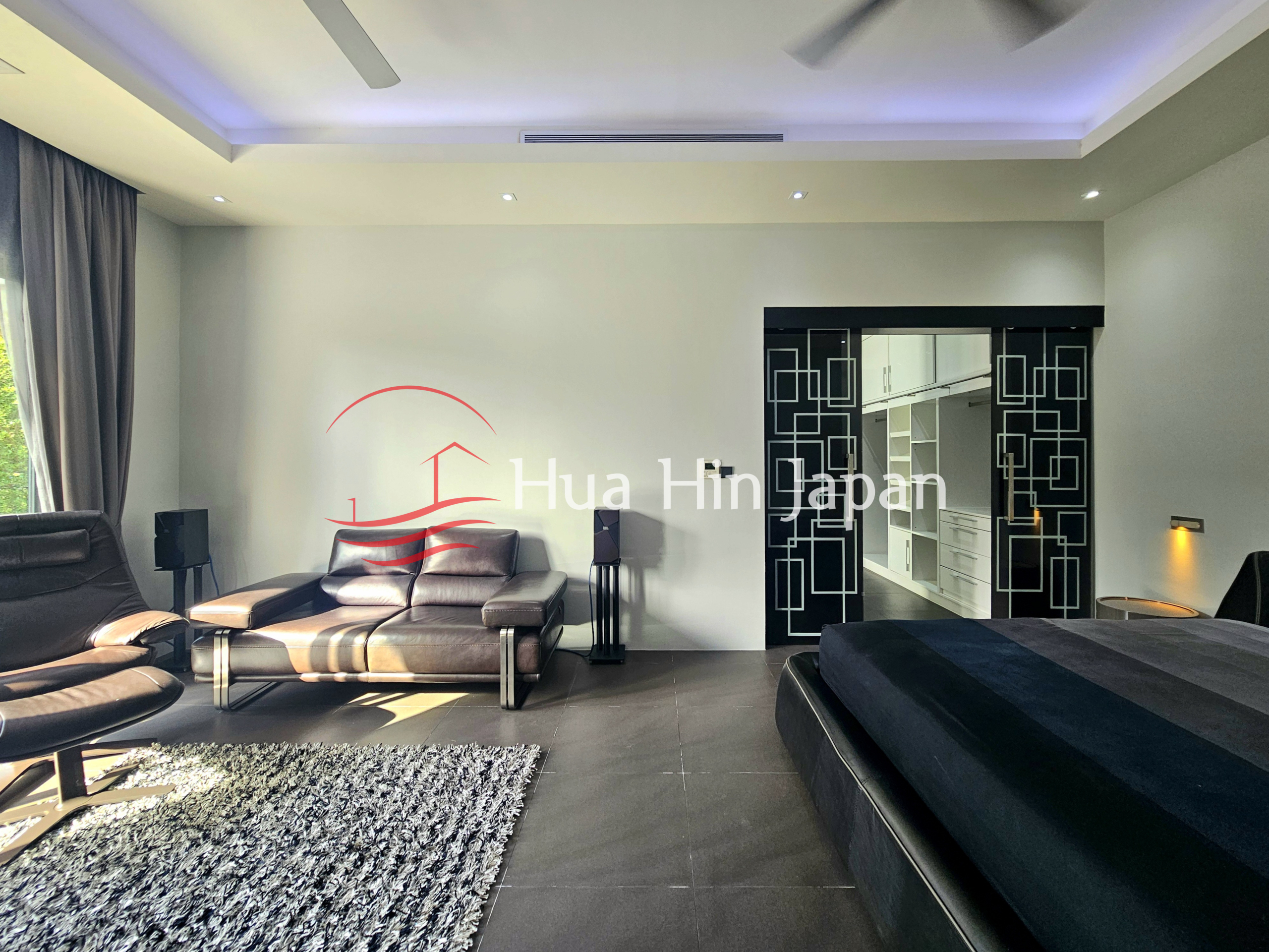 Spacious modern bedroom with a black bed, leather seating, large windows, and walk-in wardrobe