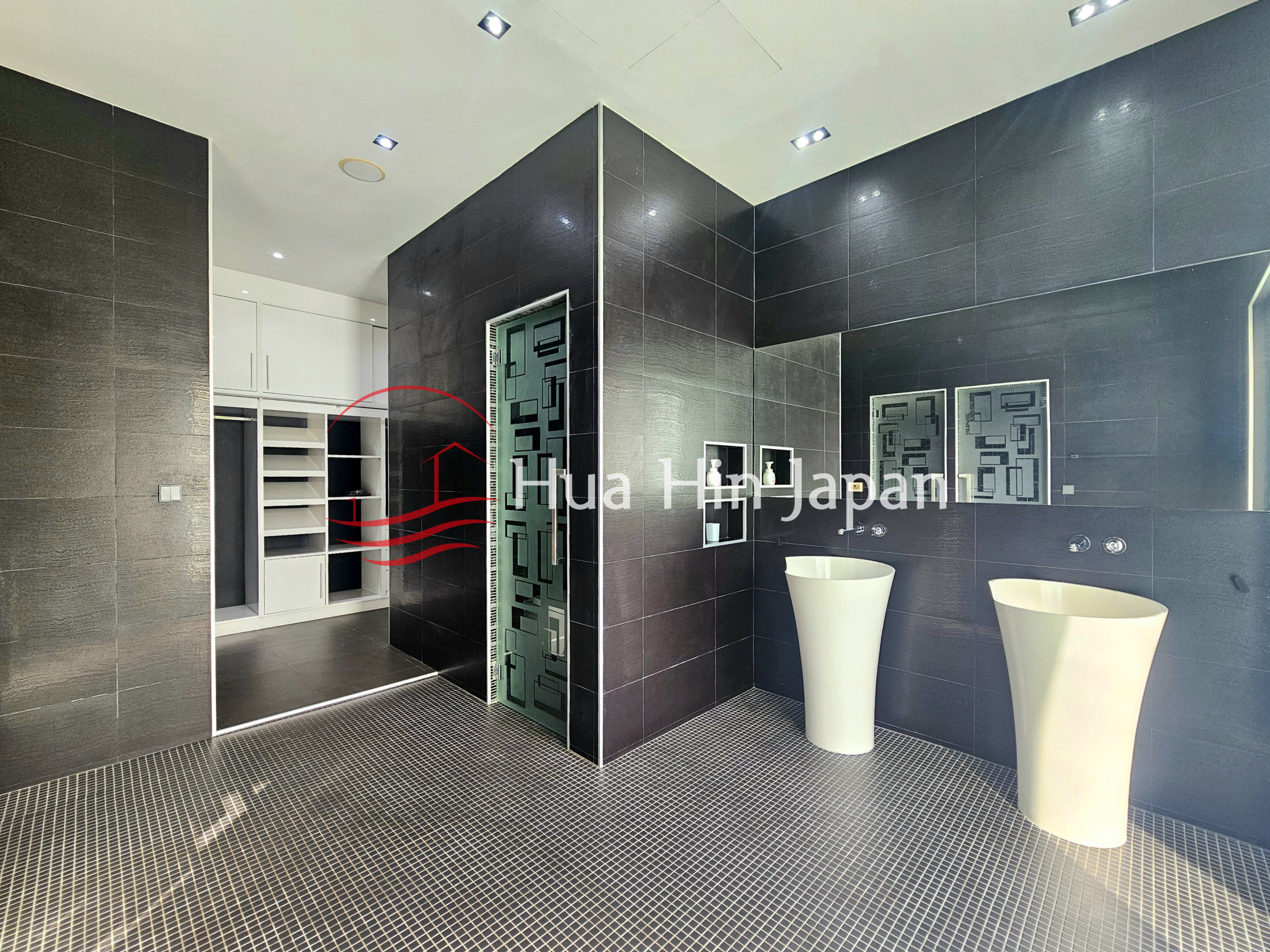 Luxurious modern bathroom with black tiled walls, mosaic flooring, twin white pedestal sinks, and a geometric glass door