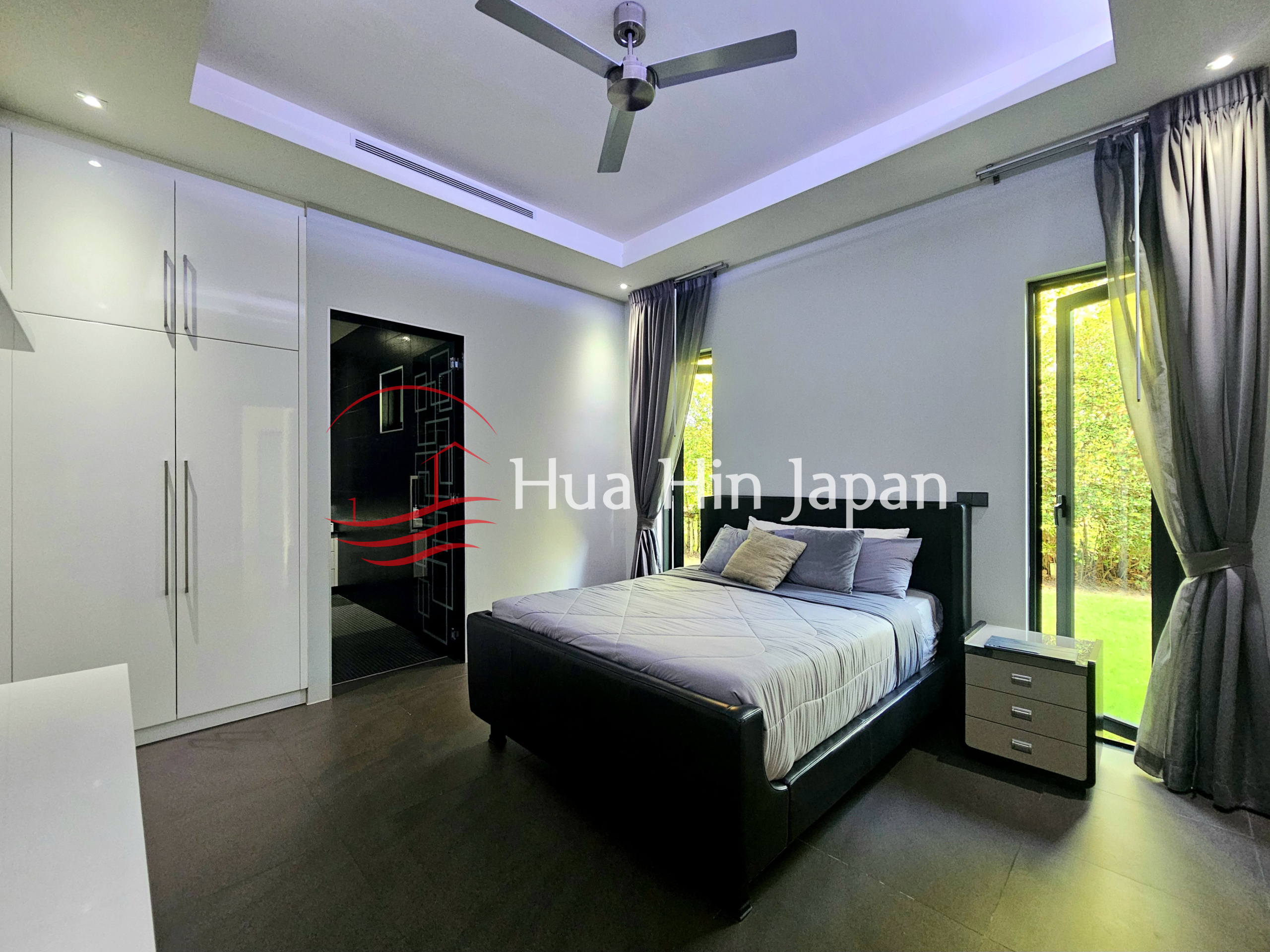 Modern bedroom with a black upholstered bed, gray bedding, white built-in wardrobes, dark flooring, ceiling fan, and large windows overlooking greenery
