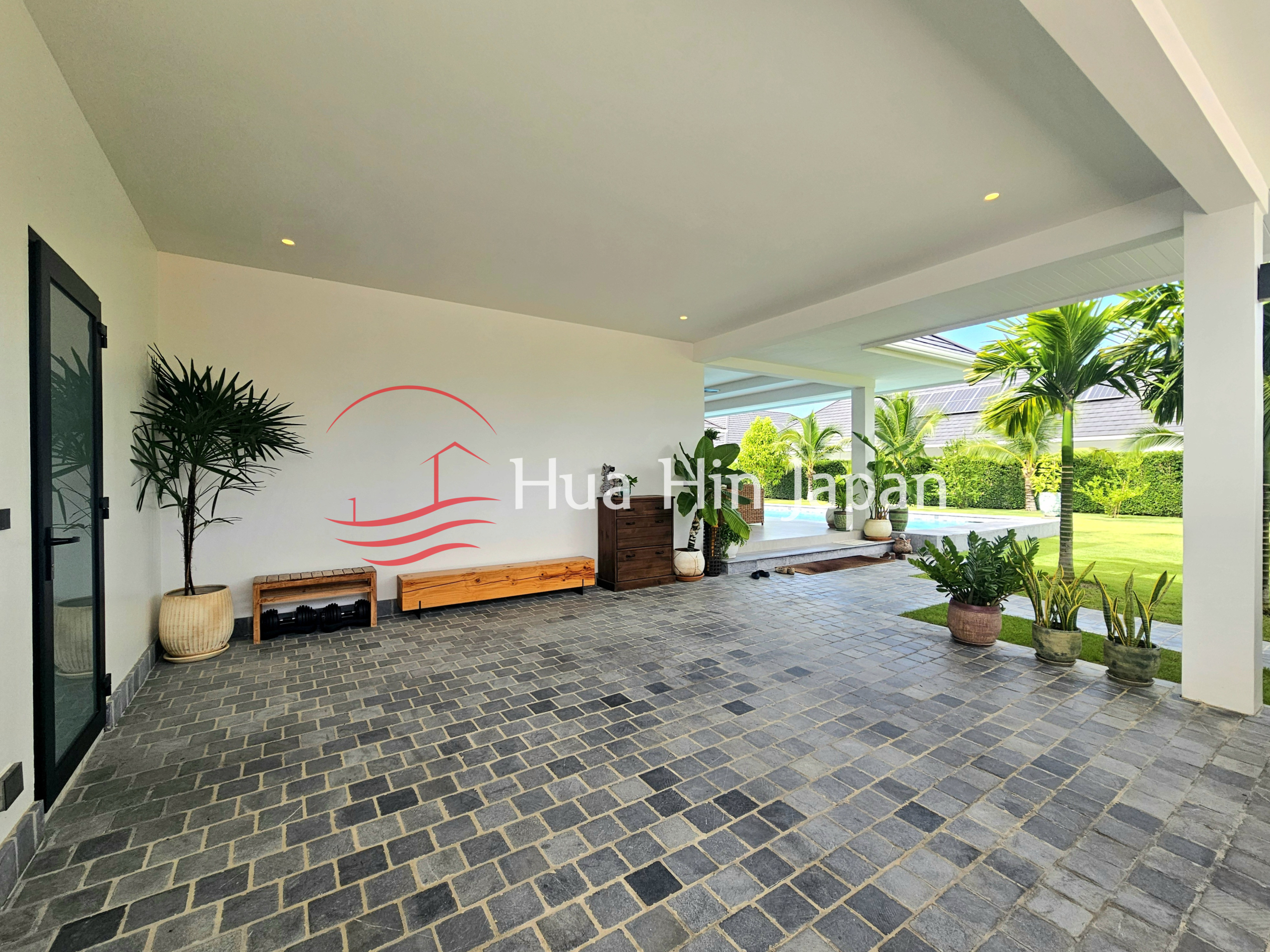 Covered outdoor patio with a paved floor, wooden bench seating, potted tropical plants, and a view of the private pool and garden