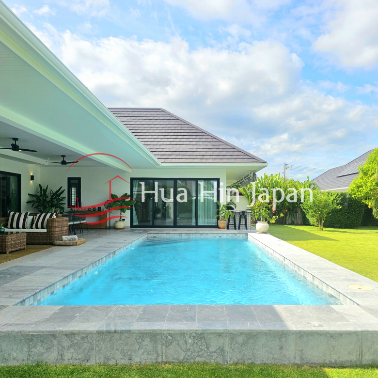 Private rectangular swimming pool beside a modern villa with a covered wicker seating area, tropical garden, and blue sky