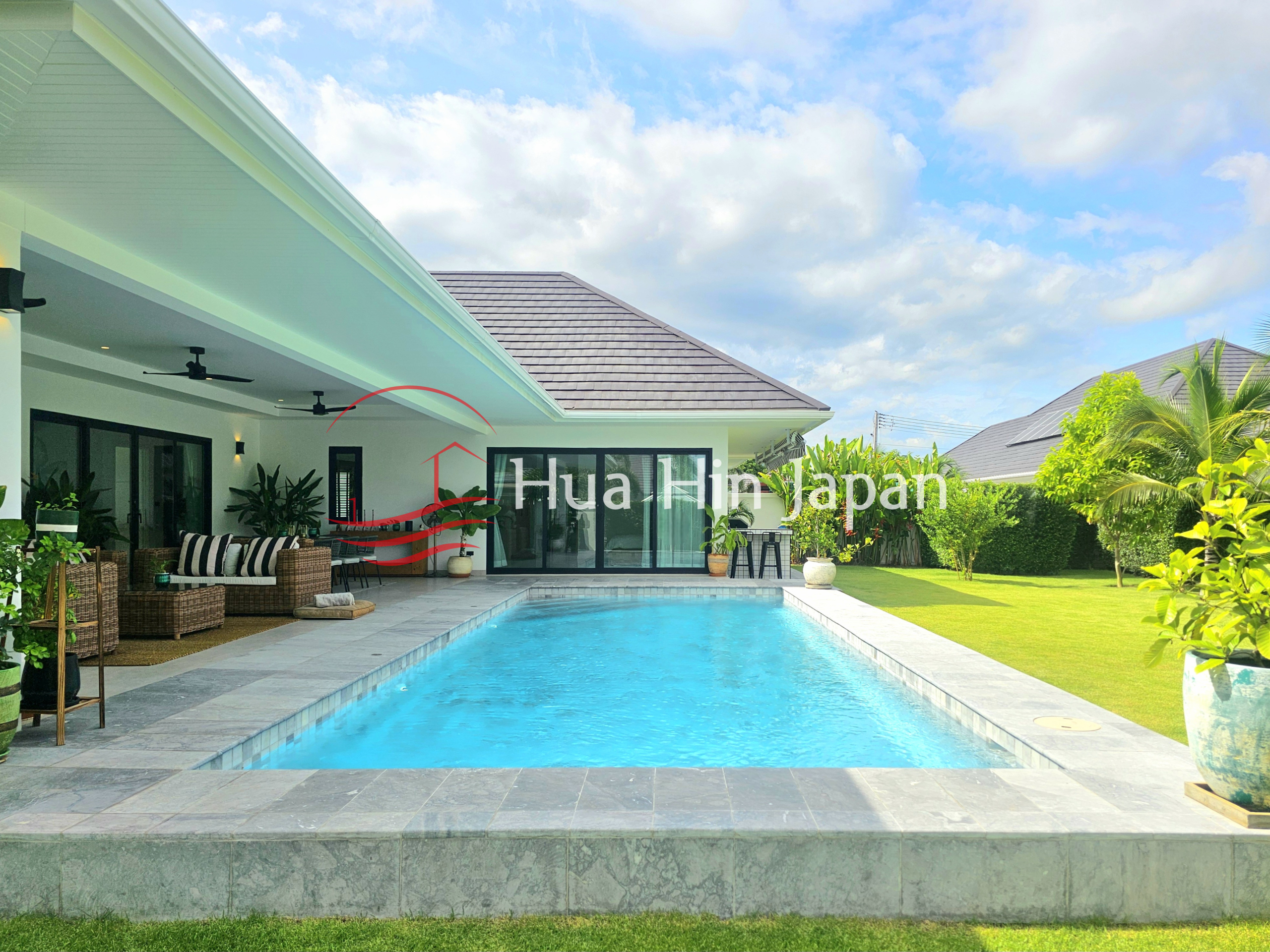 Private rectangular swimming pool beside a modern villa with a covered wicker seating area, tropical garden, and blue sky