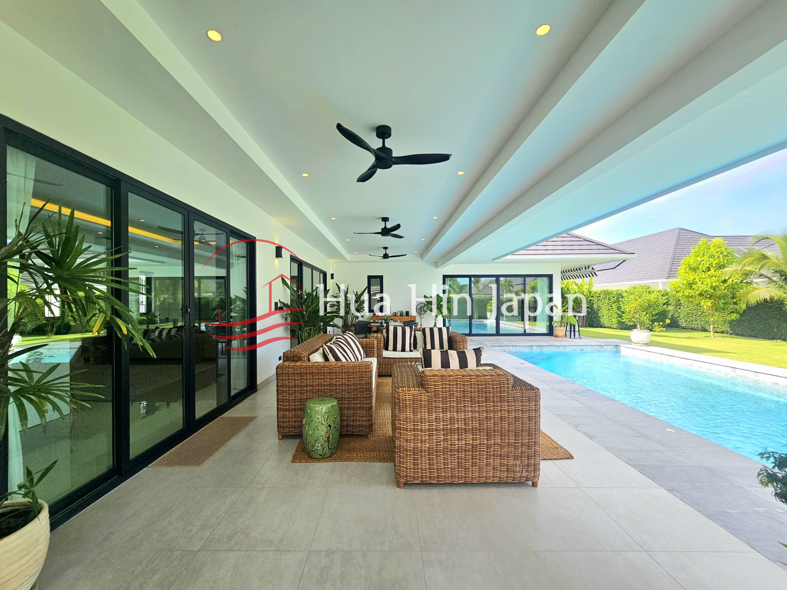 Covered outdoor lounge beside a private swimming pool with wicker seating, ceiling fans, glass doors, and a landscaped garden
