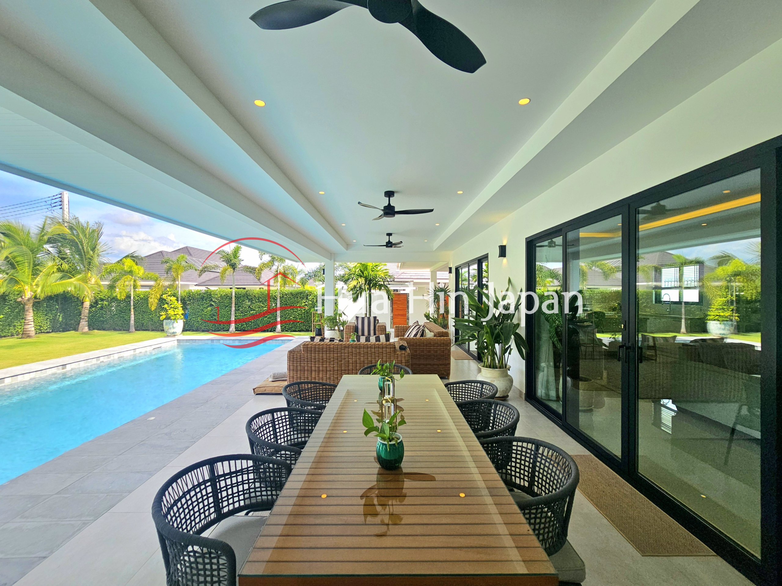 Covered poolside dining area with a long wooden table, black woven chairs, wicker seating, and tropical garden views