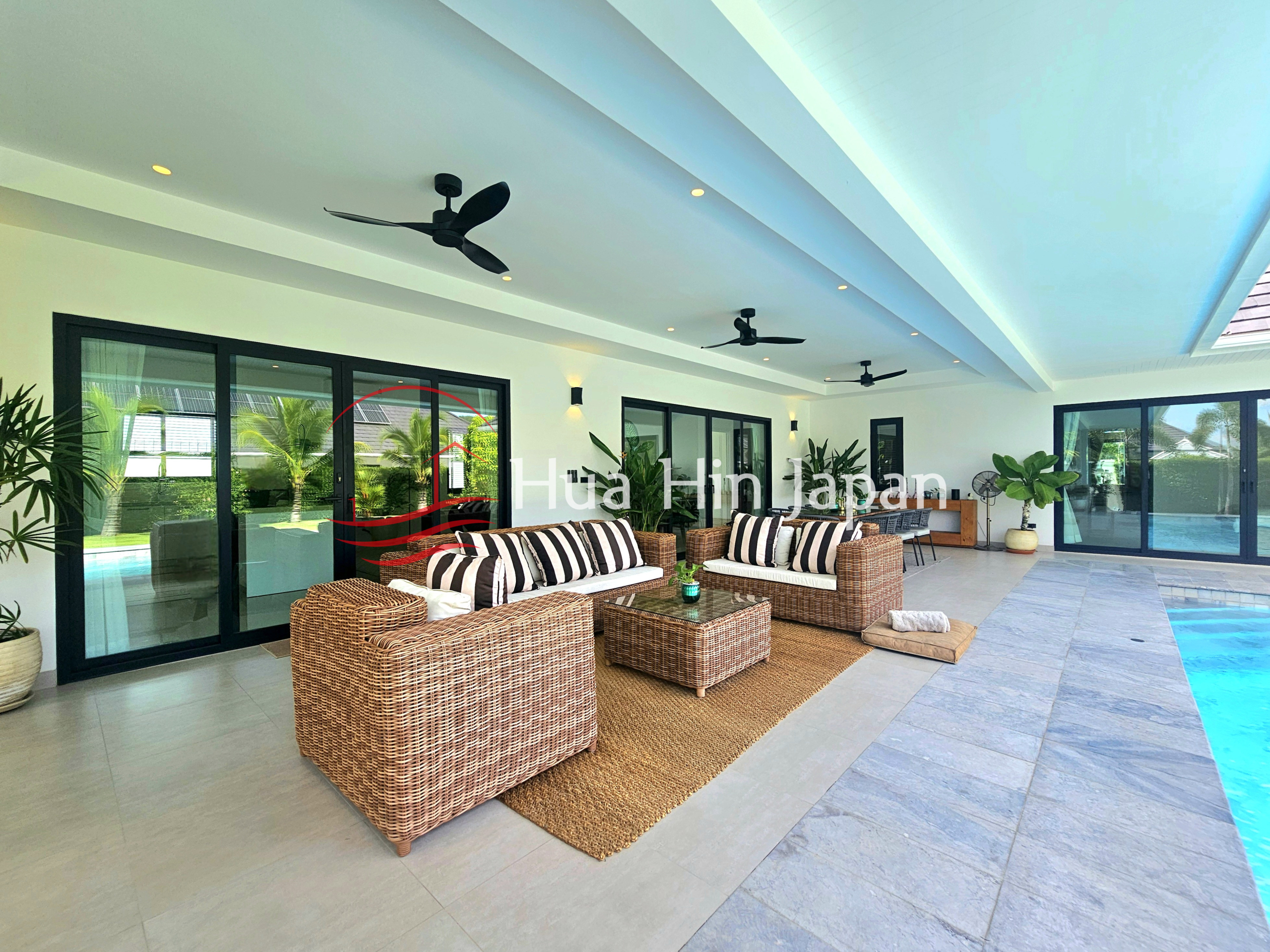 Spacious covered outdoor lounge beside a swimming pool with wicker sofas, striped cushions, ceiling fans, and glass doors