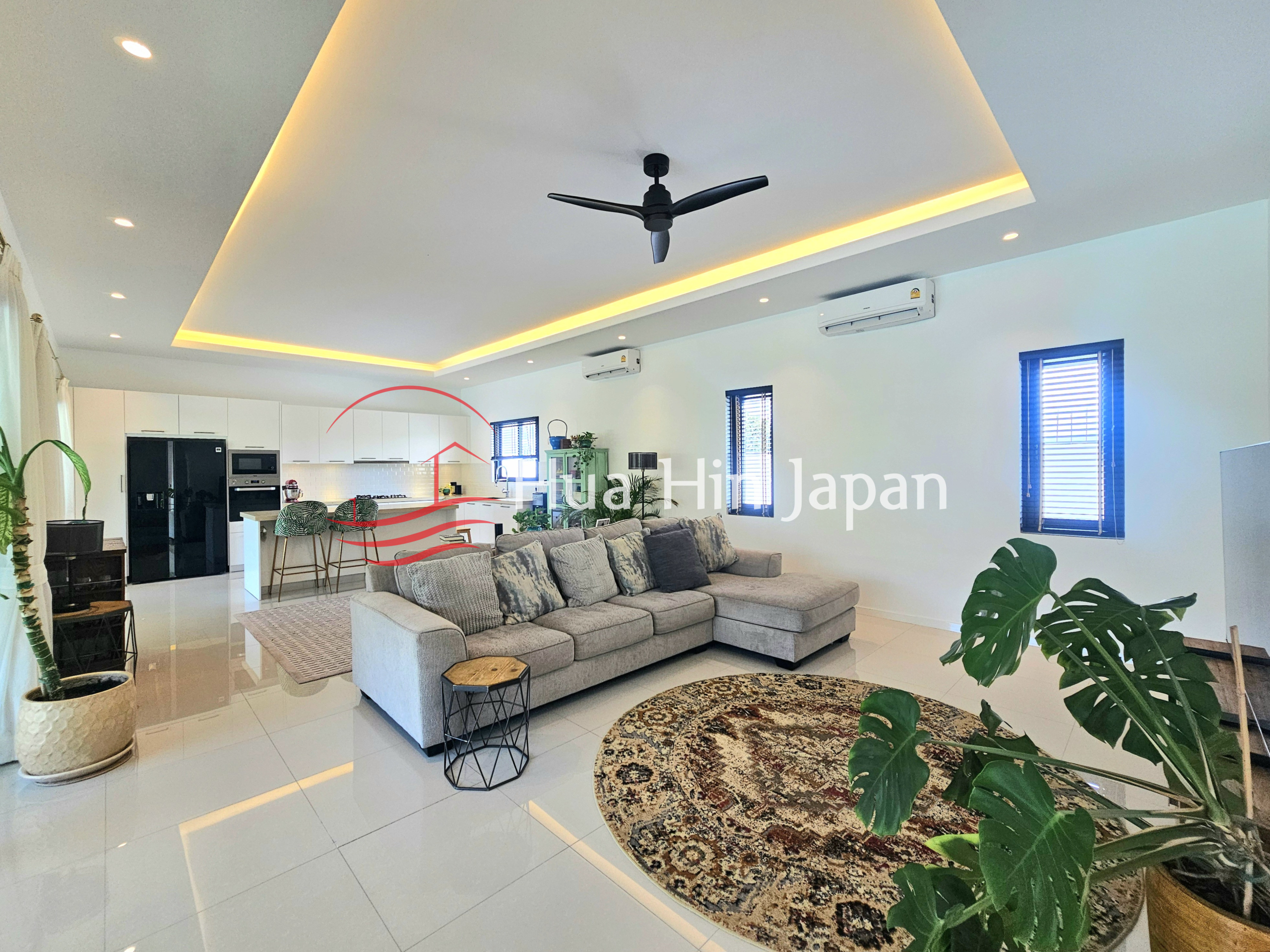Spacious modern living room with an L-shaped sectional sofa, round rug, indoor plants, ceiling fan, and open kitchen