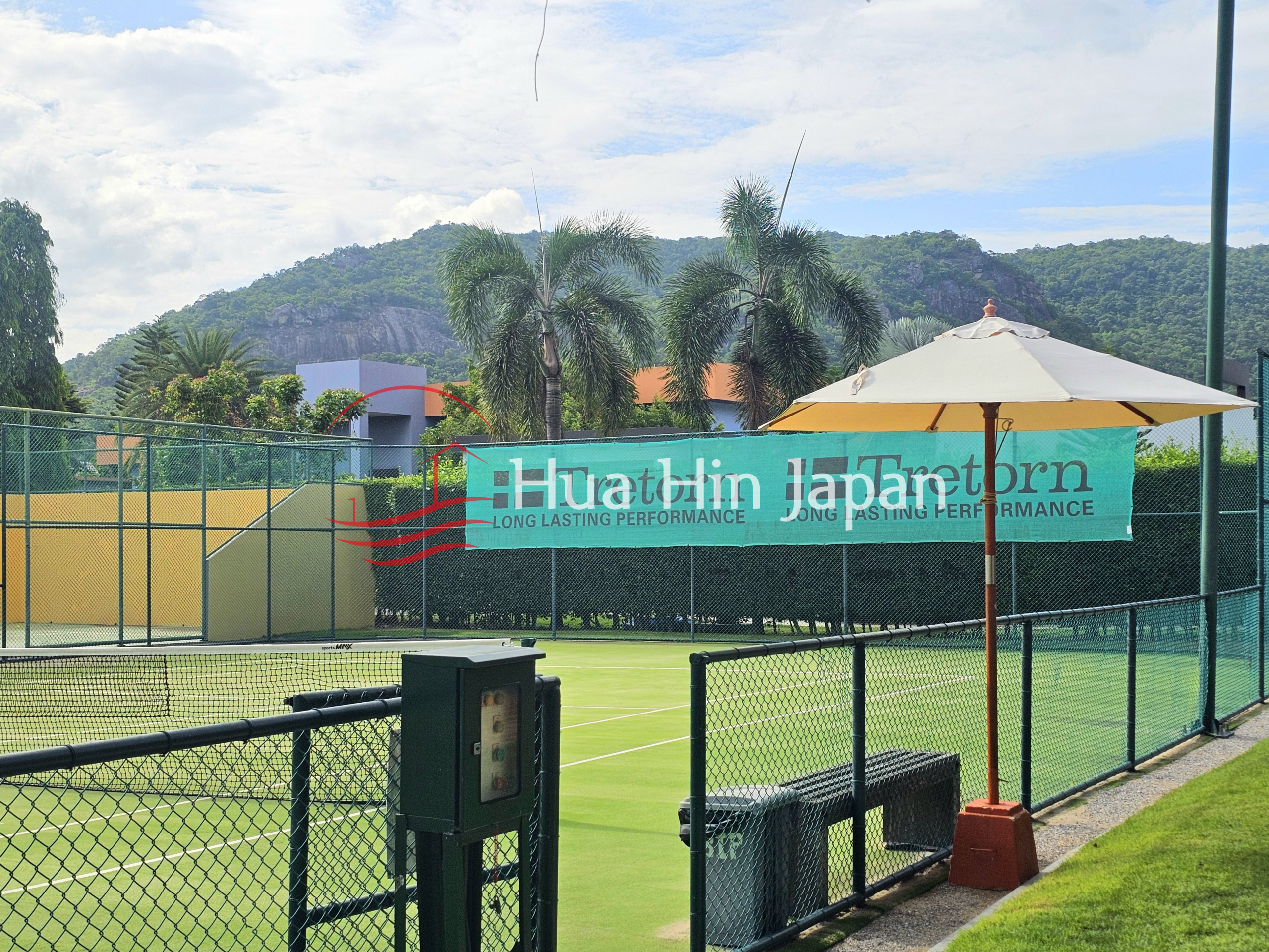 Outdoor tennis court with green fencing, a shaded seating area, and mountains in the background