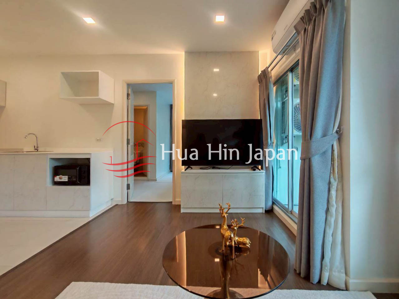 Modern living room with a TV, round coffee table, beige sofa, large windows, and compact open kitchen