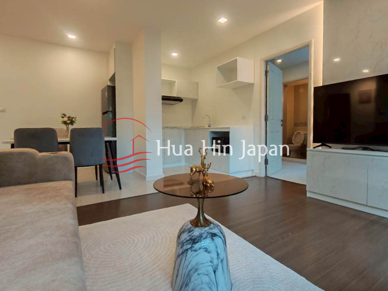 Modern apartment living room with a beige sofa, round coffee table, TV console, dining area, and open kitchen