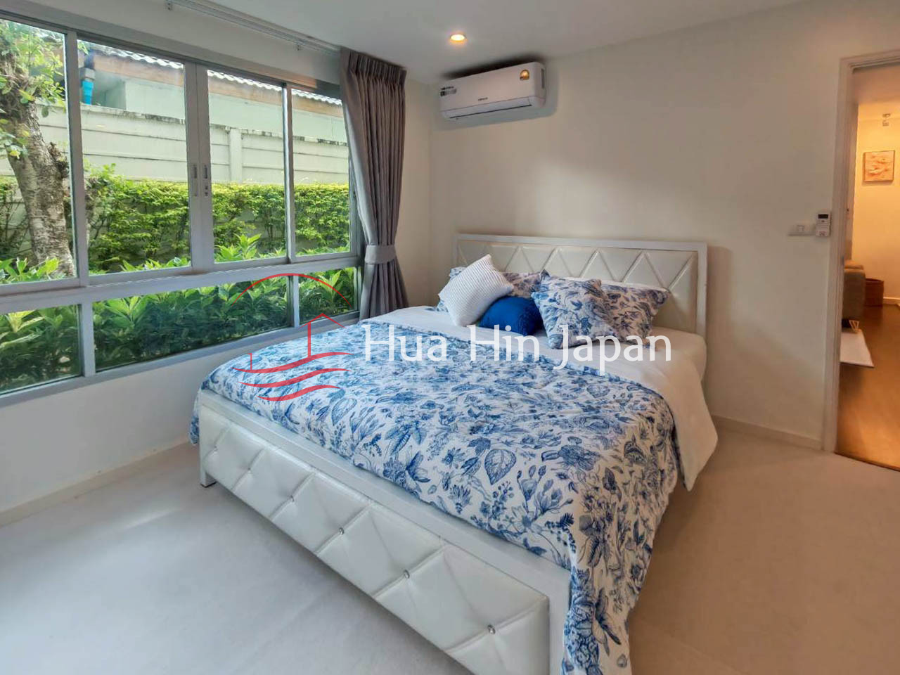 Bright modern bedroom with a white bed, blue floral bedding, large windows, and air conditioning